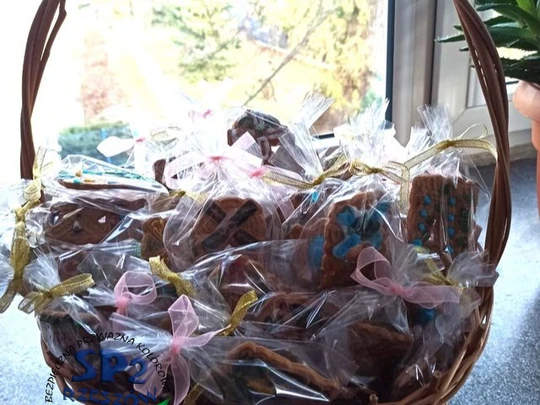 Koszyk z wikliny, a w nim zapakowane w woreczki celofanowe ozdobione pierniczki i związane kolorowymi kokardkami.