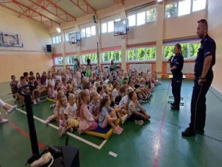 Uczniowie klas młodszych siedzą na sali gimnastycznej - spotkanie z policją
