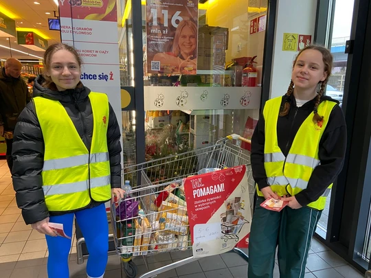 Uczennice klasy 6 w kamizelkach odblaskowych, przy wózku z ofiarowanymi produktami i rozdające ulotki akcji Caritas "Tak, Pomagam"