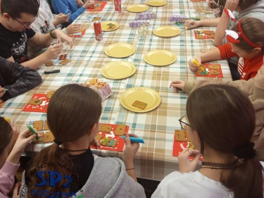 Uczniowie klasy 6 siedzą w sali przy wsp&oacute;lnym stole. Każdy ozdabia pisakami ułożone na świątecznych serwatkach pierniczki. Na środku stołu leżą puste złote papierowe talerzyki.
