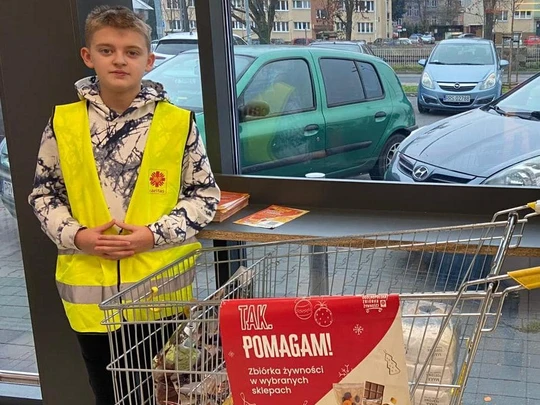 Uczeń klasy 8b w kamizelce odblaskowej, przy wózku z ofiarowanymi produktami w akcji Caritas "Tak, Pomagam"