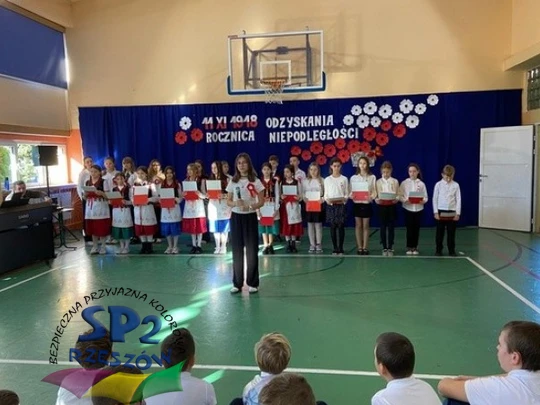 W sali gimnastycznej dzieci śpiewają, tworząc radosną atmosferę, otoczone przez koleg&oacute;w i nauczycieli