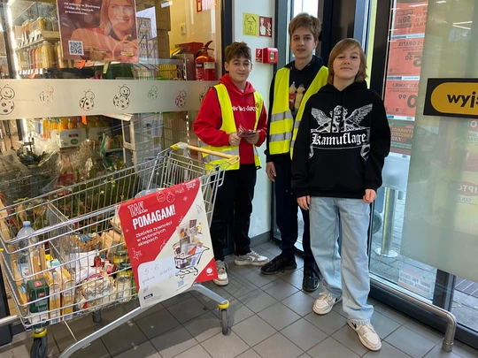 Uczniowie klasy 8b w kamizelkach odblaskowych, przy wózku z ofiarowanymi produktami i rozdający ulotki akcji Caritas "Tak, Pomagam"