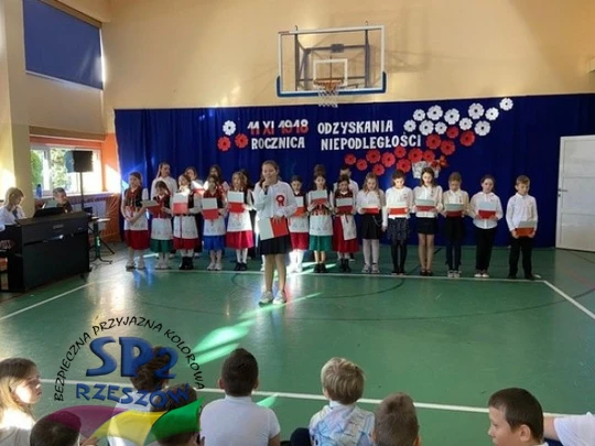 Dzieci w sali gimnastycznej śpiewają razem, uśmiechając się i trzymając się za ręce.